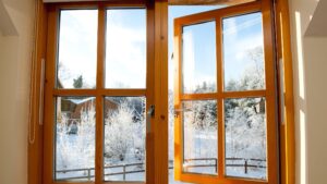 Open wooden-framed double casement windows revealing a snowy winter landscape with frosted trees, a fence, and a house under clear blue skies.