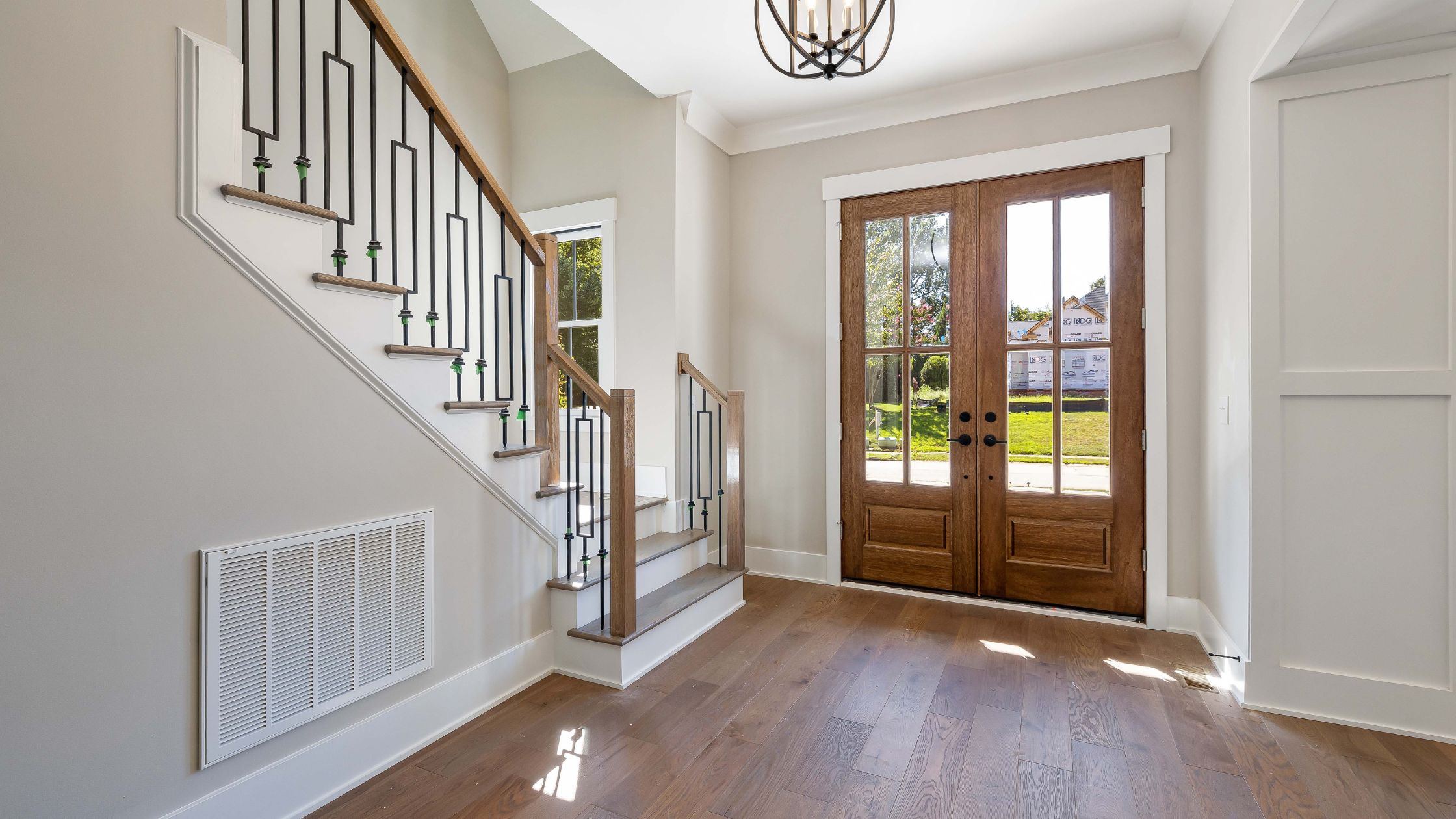 Modern home entryway featuring wooden French doors with glass panels, letting in natural light and offering a clear view of the outside, paired with a stylish staircase with black and wood railings.