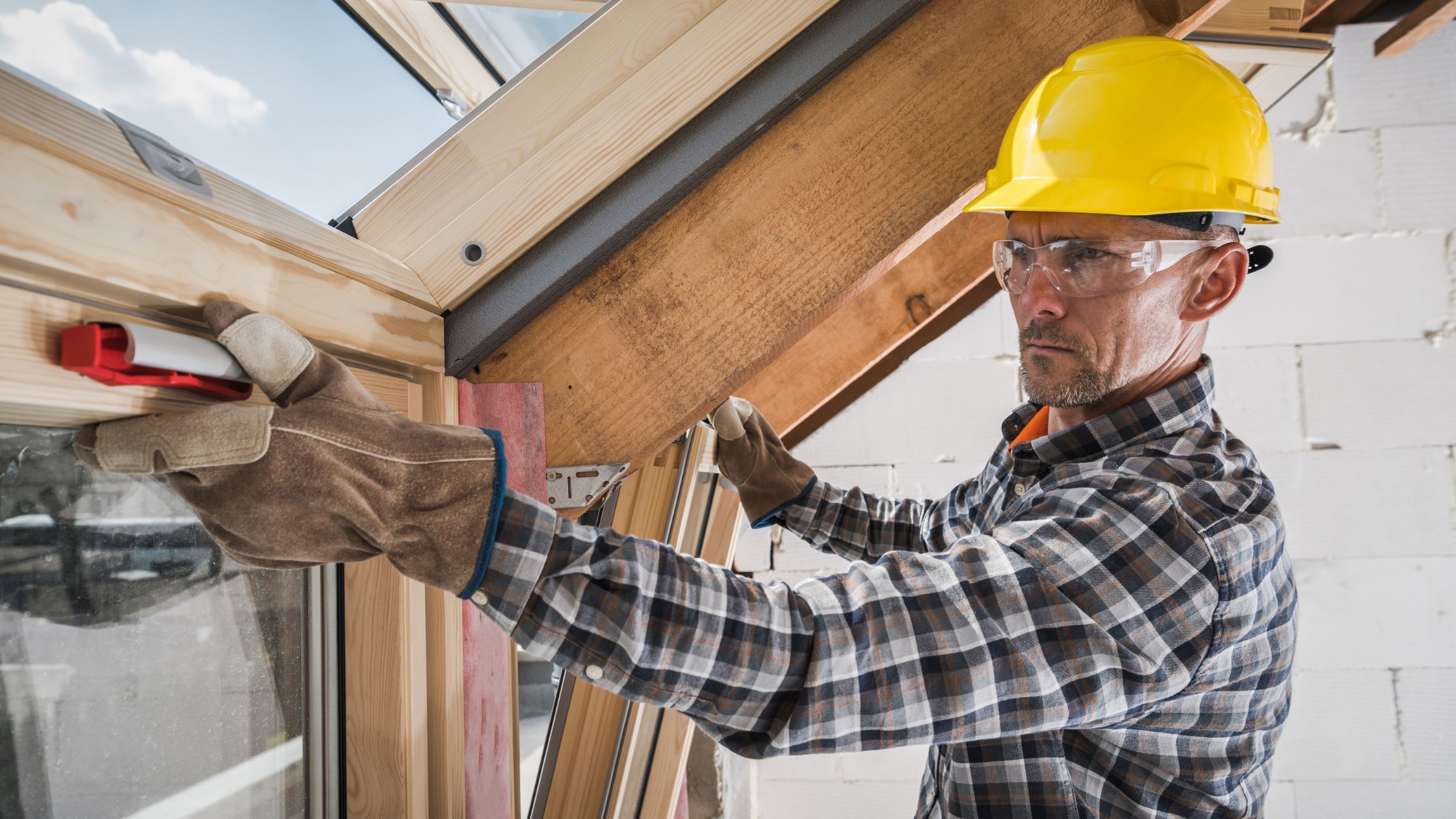 Construction worker in safety gear using a level to align a newly installed window frame on a sloped ceiling during a renovation project.