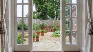 View through white French doors opening to a small backyard garden with potted plants, gravel, and a wooden fence, framed by elegant cream-colored curtains.