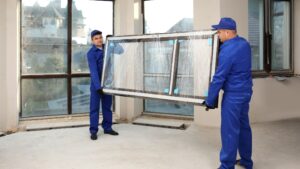 Two workers in blue uniforms carry a large, wrapped window frame inside a building under renovation, with tall glass windows in the background.