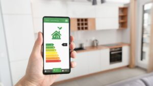 Person holding a smartphone displaying a smart home energy efficiency rating app, showing a green house icon and an A+ energy rating chart in a modern kitchen setting.