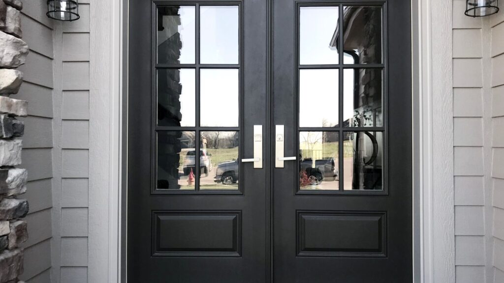 Black double steel front doors with glass panel inserts and modern silver handles, set against a light gray exterior wall with stone accents and outdoor wall sconces.