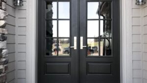 Black double steel front doors with glass panel inserts and modern silver handles, set against a light gray exterior wall with stone accents and outdoor wall sconces.