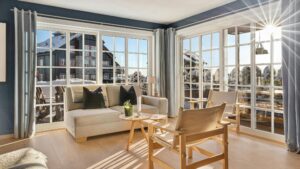 Bright and cozy living room with large, white-framed windows and glass doors letting in natural sunlight, featuring light wood flooring, a beige sofa, and minimalist wooden furniture. Snow-covered houses are visible outside.