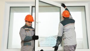 Two construction workers wearing safety helmets and uniforms installing a large white-framed window indoors, representing window replacement in a residential or commercial setting.
