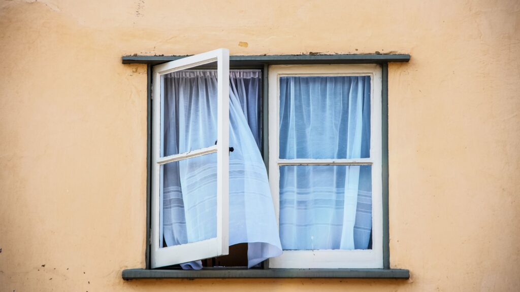 An open white casement window with sheer white curtains billowing outward, set in a light peach-colored wall of a building.