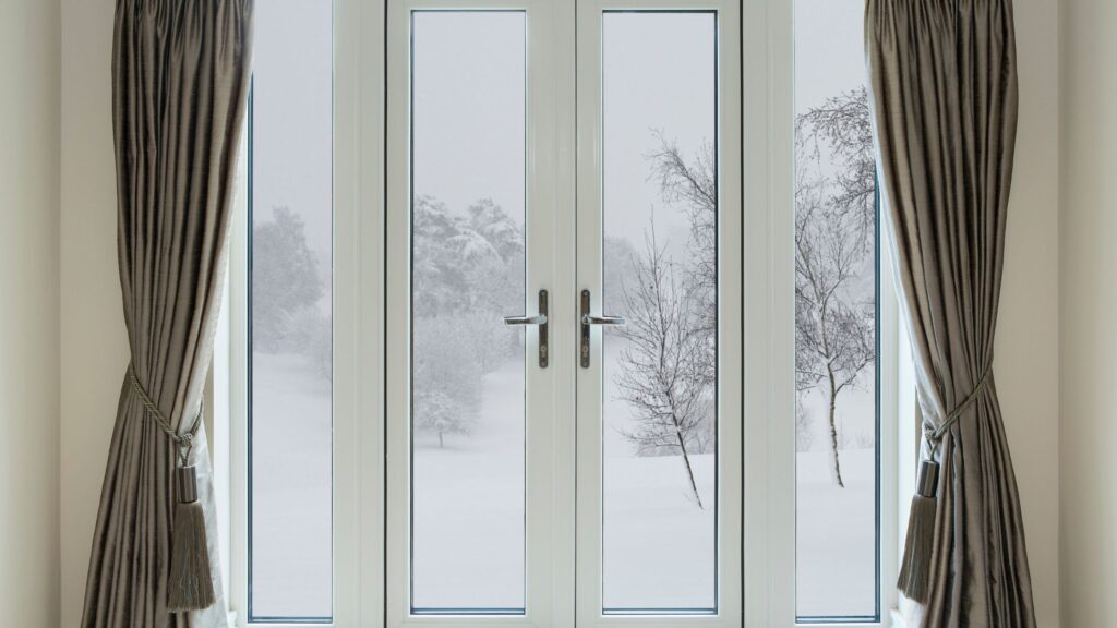 A pair of elegant French doors with large glass panels, framed by light-colored curtains tied back with tassels. The view outside shows a snowy landscape with trees, creating a serene winter atmosphere.