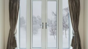 A pair of elegant French doors with large glass panels, framed by light-colored curtains tied back with tassels. The view outside shows a snowy landscape with trees, creating a serene winter atmosphere.