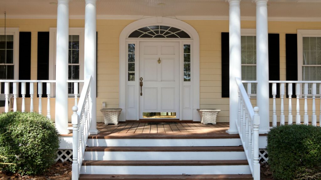 A welcoming front porch with white columns, a wooden deck, and a white door with glass panels. Two decorative planters are placed on either side of the stairs, framed by neatly trimmed bushes.