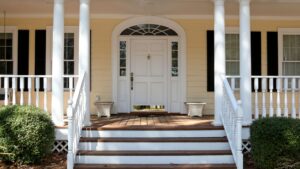 A welcoming front porch with white columns, a wooden deck, and a white door with glass panels. Two decorative planters are placed on either side of the stairs, framed by neatly trimmed bushes.