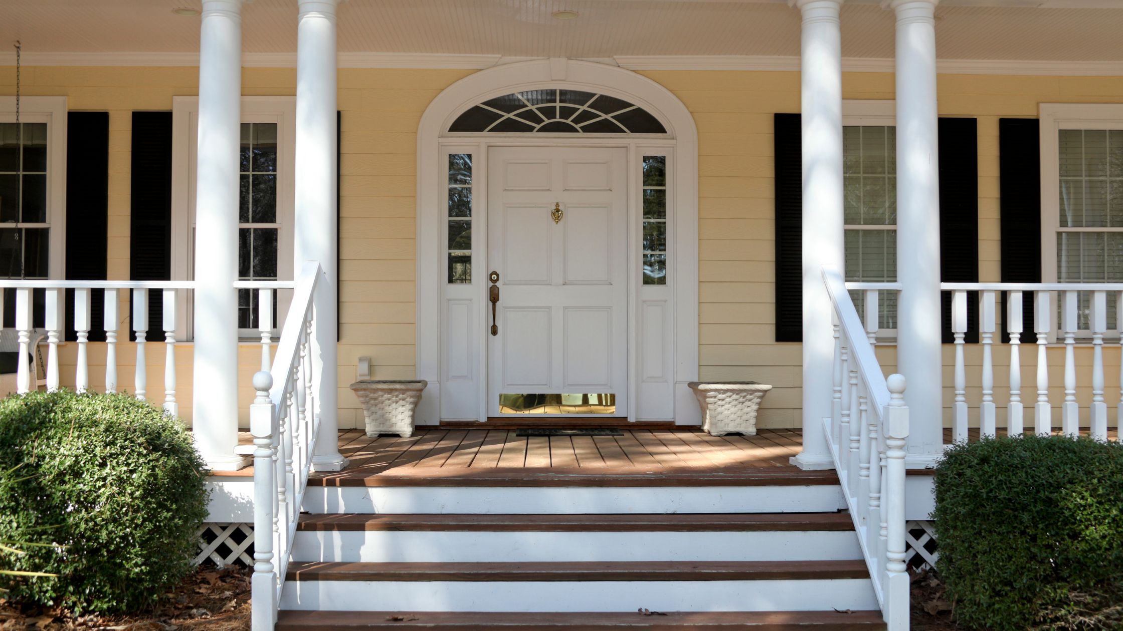 A welcoming front porch with white columns, a wooden deck, and a white door with glass panels. Two decorative planters are placed on either side of the stairs, framed by neatly trimmed bushes.