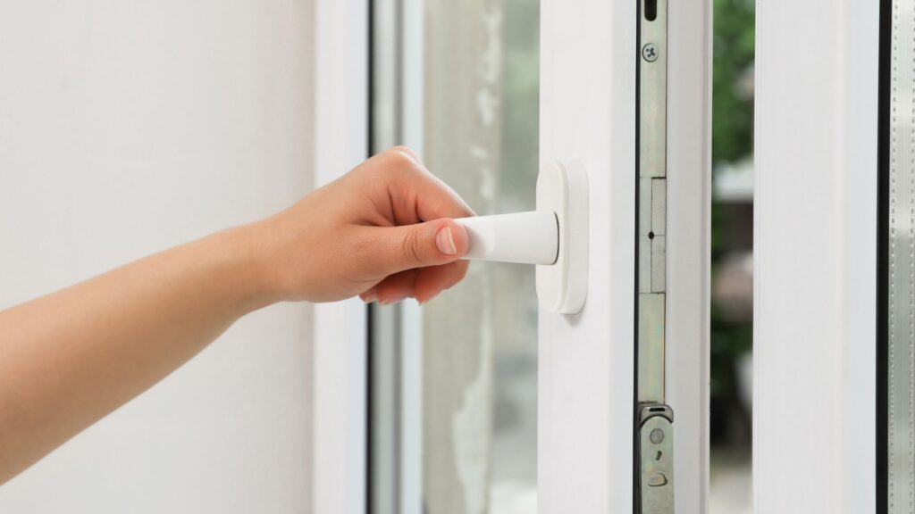 Close-up of a person's hand turning the white handle of a modern casement window, partially opened to show the window's locking mechanism and frame.