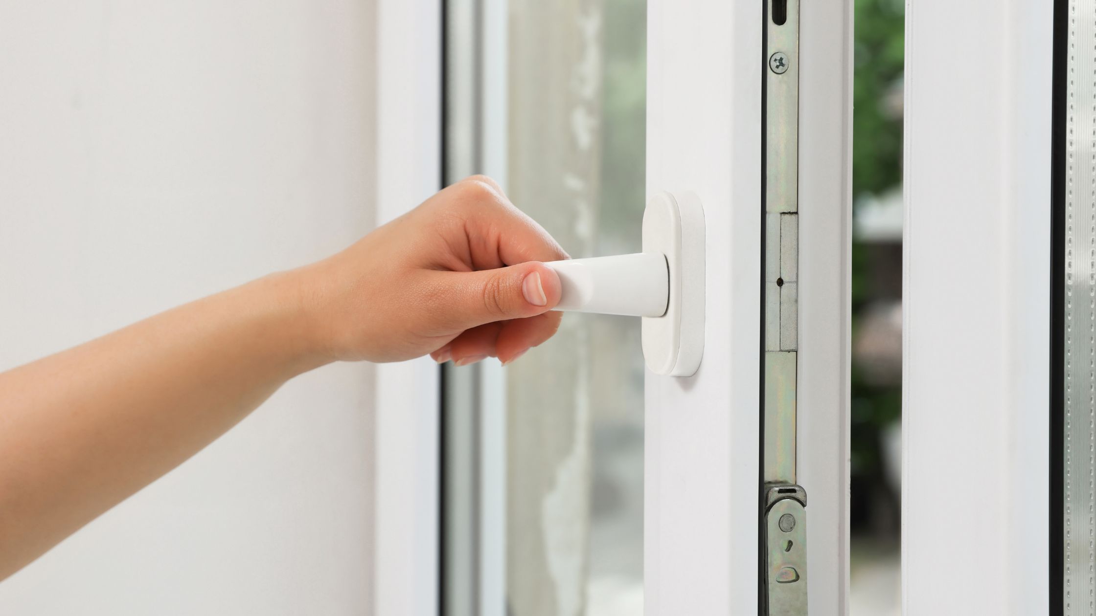 Close-up of a person's hand turning the white handle of a modern casement window, partially opened to show the window's locking mechanism and frame.