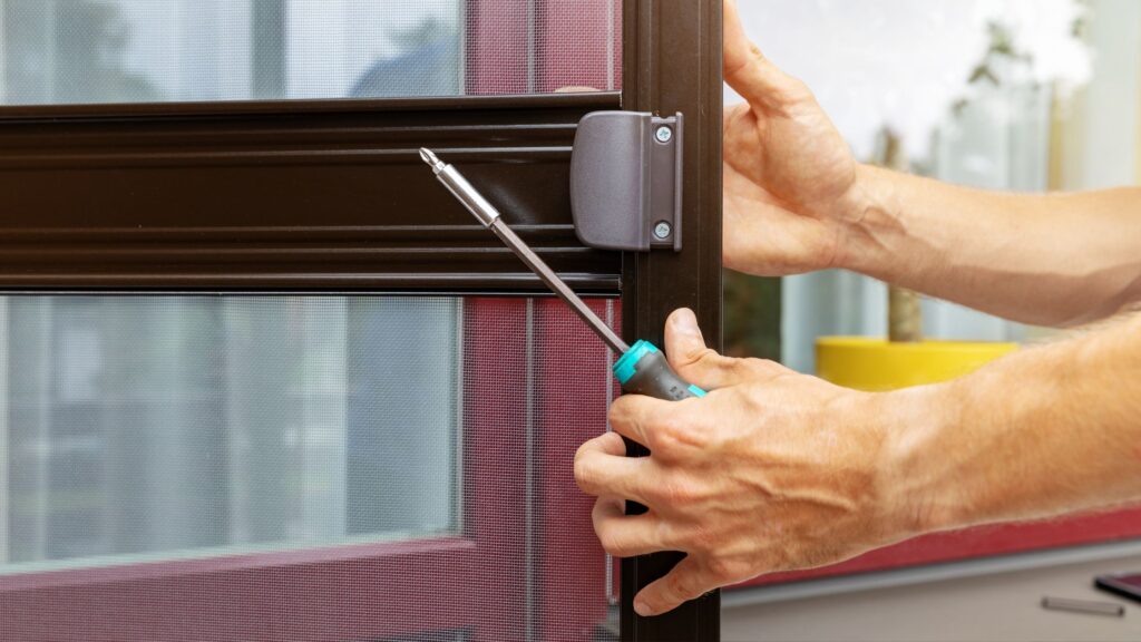 Person using a screwdriver to remove or install a latch on a brown-framed screen door, with mesh and window in the background.
