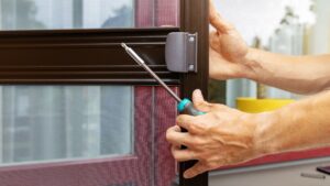 Person using a screwdriver to remove or install a latch on a brown-framed screen door, with mesh and window in the background.