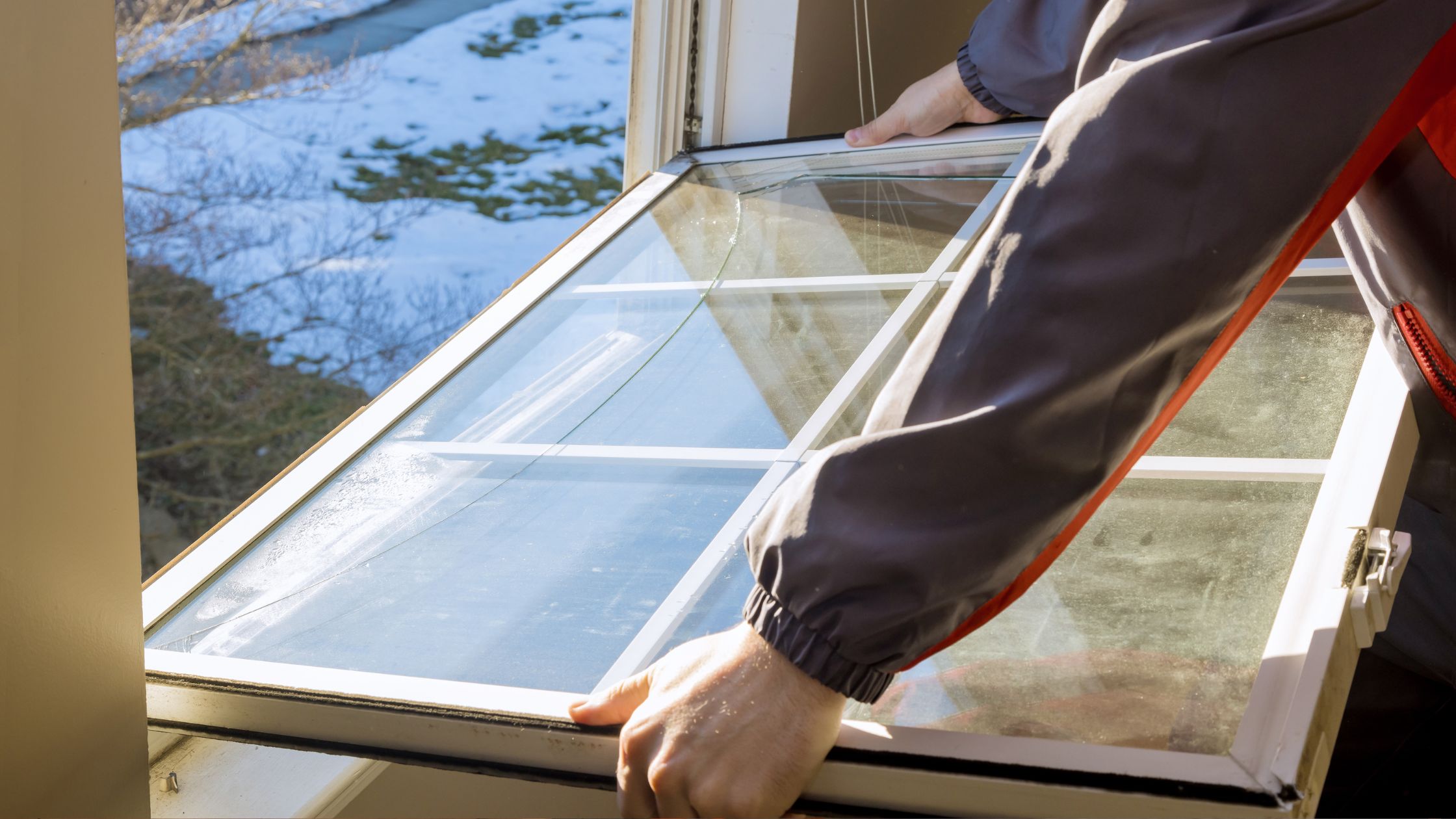 Worker removing a cracked window sash during replacement; snow and trees visible outside through the open window.