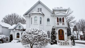 Snow-covered Victorian-style white brick house with arched windows, wooden double front door, balcony, and surrounding winter landscape.