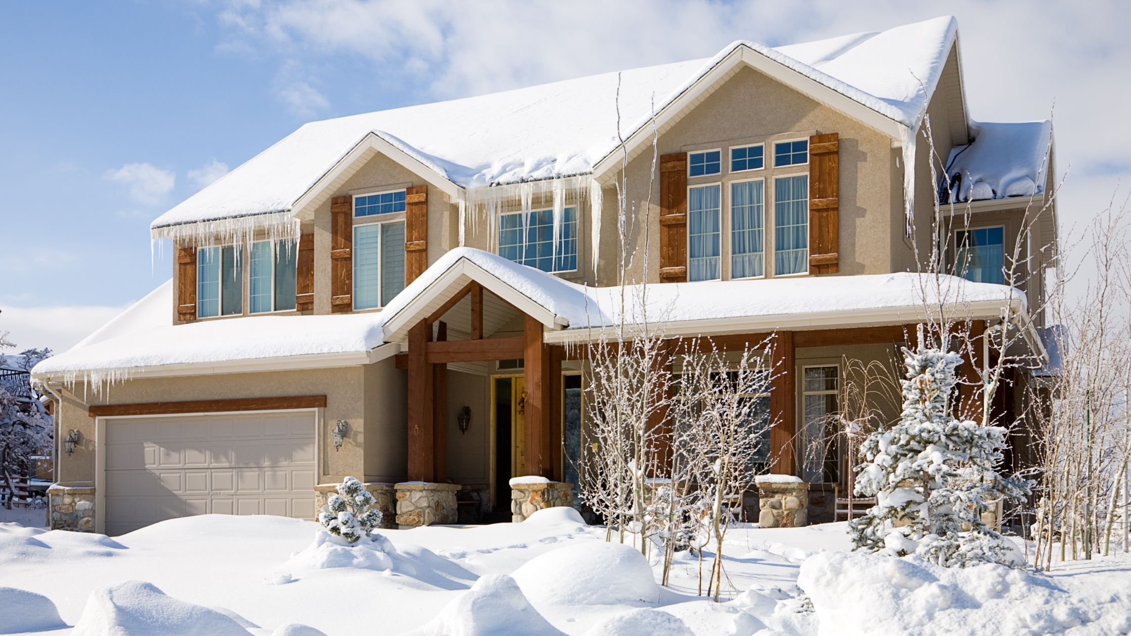 Modern two-story home with wooden accents and large windows, surrounded by deep snow and icicles hanging from the roof on a sunny winter day.