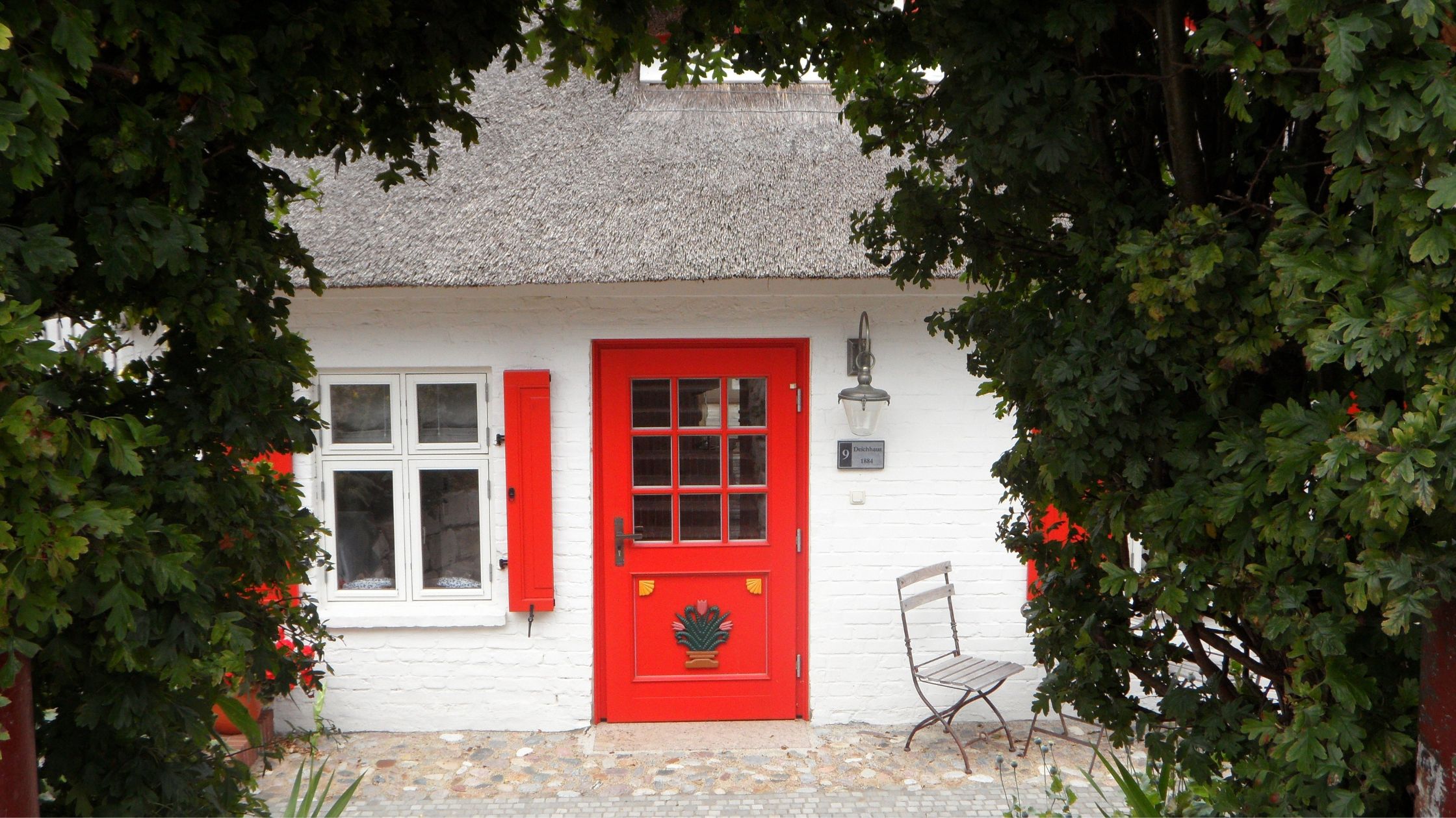 Front view of a charming white brick house with a thatched roof, featuring a bright red front door with floral design and matching red window shutters, framed by green foliage.