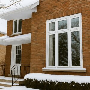 Exterior view of a brick house in winter with large white-framed energy-efficient triple-pane windows and snow-covered ground and roof edges.