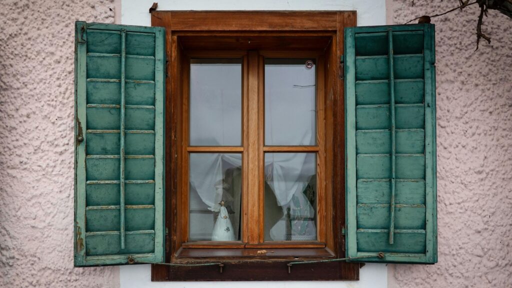 Common window problems in Winnipeg homes shown by an old wooden window with peeling paint, damaged shutters, and visible wear from harsh weather.